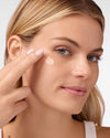 Woman applying COOLA Sunless Tan Anti-Aging Face Moisturizer to cheek with white background.