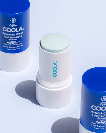 Three COOLA Refreshing Water Hydration Sticks sitting on blue surface with one uncapped