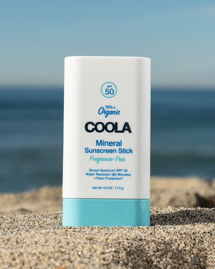 COOLA Mineral Sunscreen Stick Fragrance-Free SPF 50 sitting in the sand with scenic ocean background