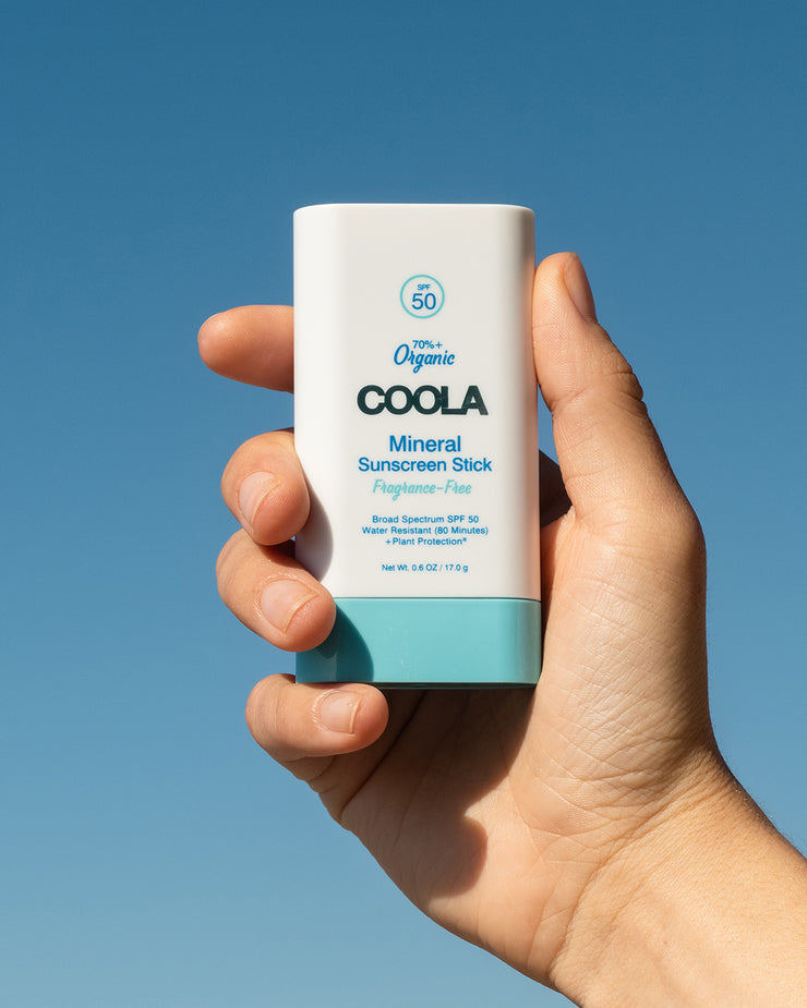 COOLA Mineral Sunscreen Stick Fragrance-Free SPF 50 being held in hand with a blue sky background.