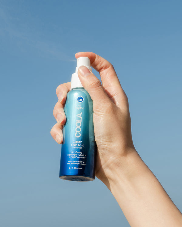 COOLA Classic Face Mist Sunscreen SPF 50 being held in hand spraying in the air with a blue sky background.