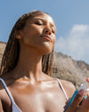 Woman applying COOLA Classic Face Mist Sunscreen to face with blue sky and coastal background