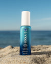 Bottle of COOLA Classic Face Mist sunscreen SPF 50 sitting in sand with a scenic ocean background.