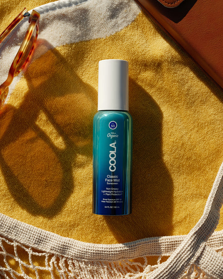 Bottle of COOLA Classic Face Mist sunscreen SPF 50 laying on yellow towel with mesh bag and sunglasses at the beach.