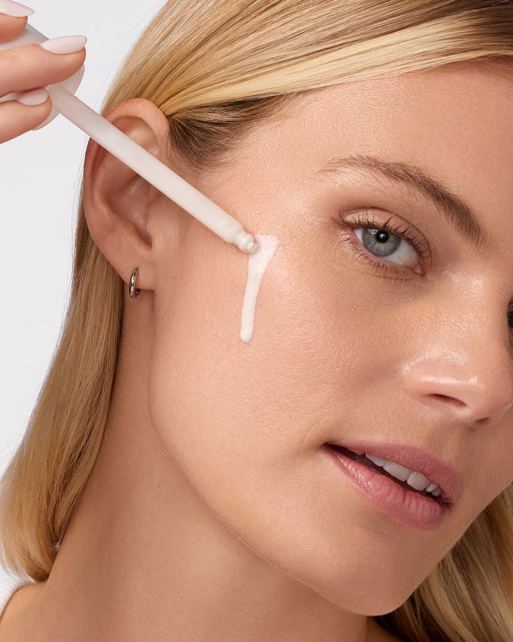 Close-up of woman applying COOLA Sun Silk Drops Sunscreen SPF 30 to cheek with white background