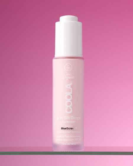 Bottle of COOLA Sun Silk Drops Sunscreen SPF 30 sitting on glass shelf against pink background