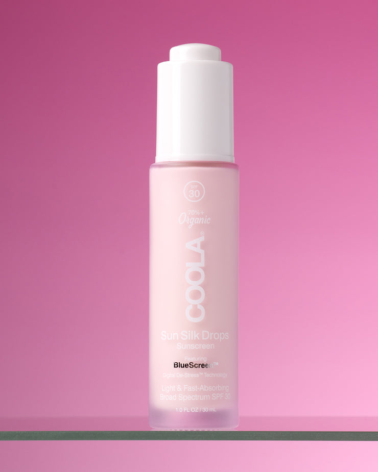Bottle of COOLA Sun Silk Drops Sunscreen SPF 30 sitting on glass shelf against pink background