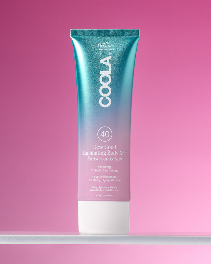 Tube of COOLA Dew Good Illuminating Body Melt Lotion SPF 40 sitting on glass shelf with pink background