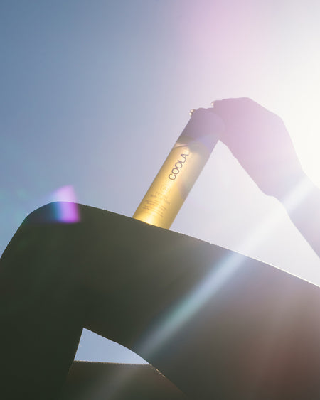 Person holding COOLA Body Oil Mist with bright sunny sky background