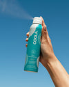 Hand holding a COOLA sunscreen spray can against a clear blue sky