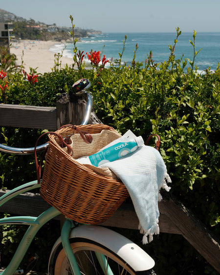 Bicycle with a basket containing a towel and a bottle, set against a scenic beach backdrop.