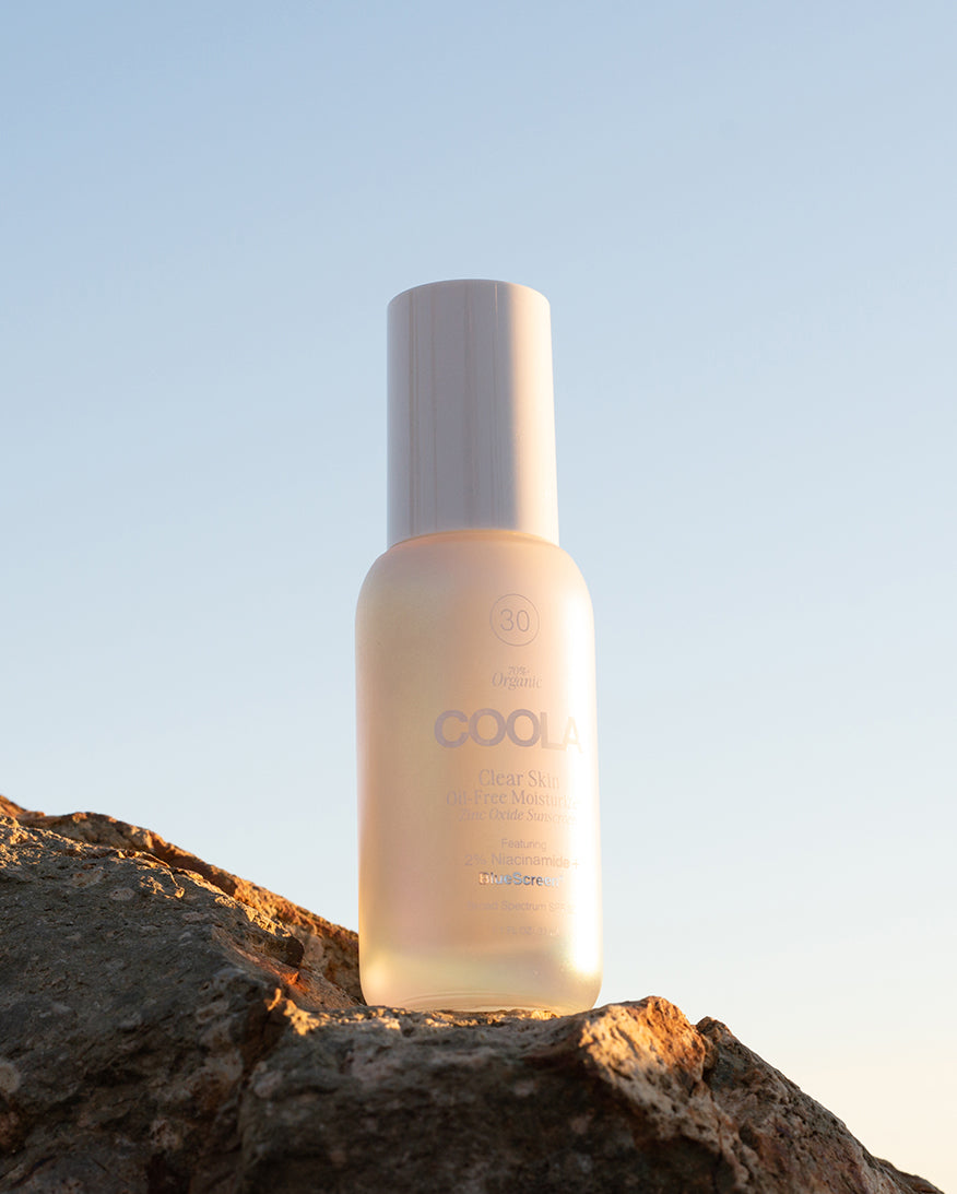 White bottle of COOLA Clear Skin Oil-Free Moisturizer Zinc Oxide Sunscreen sitting on rock with blue sky background.