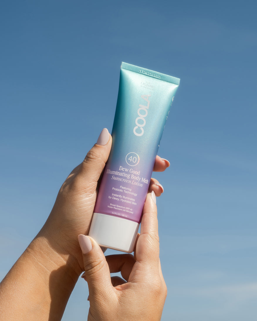 Hands holding tube of COOLA Dew Good Illuminating Body Melt Lotion SPF 40 with blue sky background.