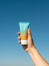 Travel-sized body lotion sunscreen with orange gradient being held by a person up to a blue sky
