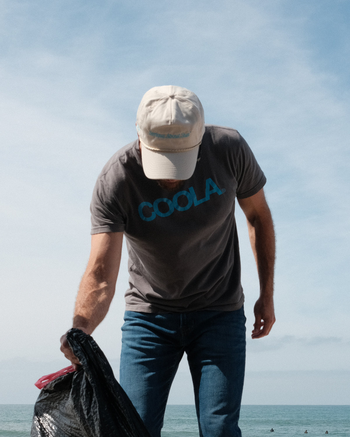 Person wearing a beige cap and COOLA t-shirt picking up trash near the ocean, holding a black trash bag under a partly cloudy sky