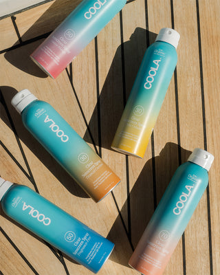 Collection of five different Clear Sunscreen Sprays laying down on wooden deck.