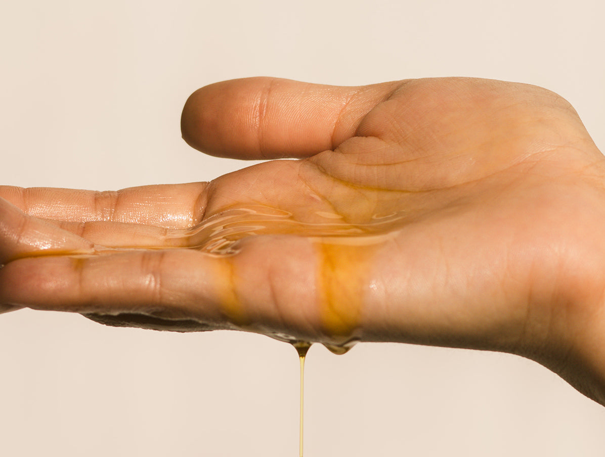 Close-up of a persons hand with yellow oil sunscreen dripping over hand with a white backdrop