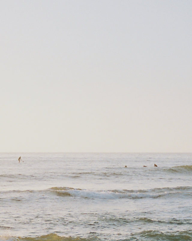 A calm ocean scene under a clear sky, with gentle waves and a few surfers in the distance, one standing on a paddleboard and others sitting on their boards.