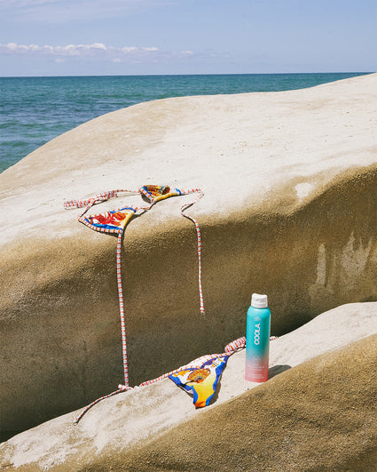 Colorful bikini and a turquoise bottle of COOLA sunscreen placed on smooth tan rocks by the ocean under a clear blue sky.