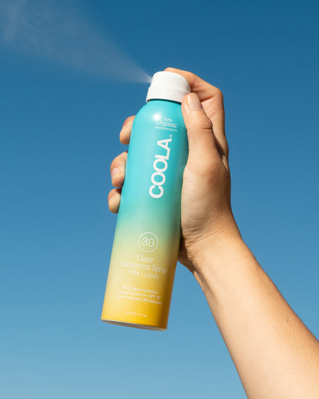 Clear Sunscreen Spray, Pina Colada fragrance in a human hand against a blue sky.