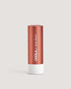 A stick of COOLA Liplux Tinted Lip Balm in a nude beach shade, with a copper-colored body and white base.