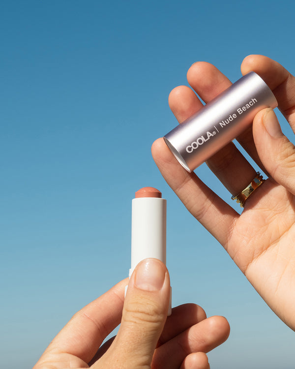 COOLA Nude Beach Mineral Liplux with cap taken off showing formula against sky blue background.