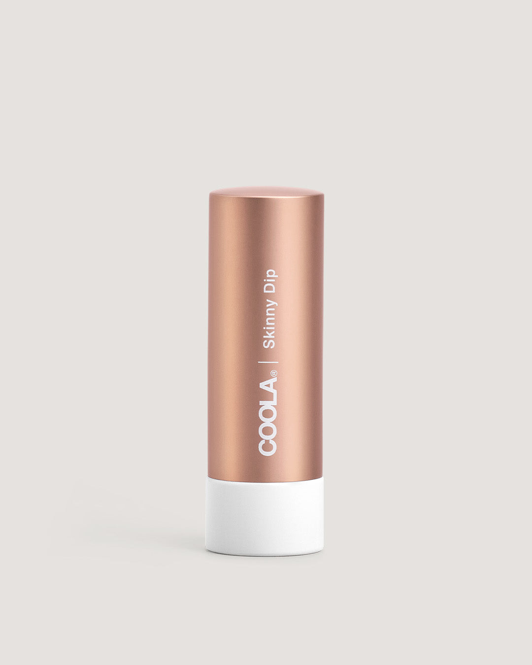 A stick of COOLA Liplux Tinted Lip Balm in a rose gold colored tube with the label 'Skinny Dip' on the front.