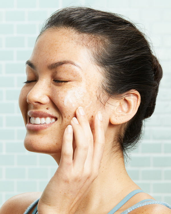 Woman applying Coola Glowing Greens Detoxifying Facial Cleansing Gel in bathroom.