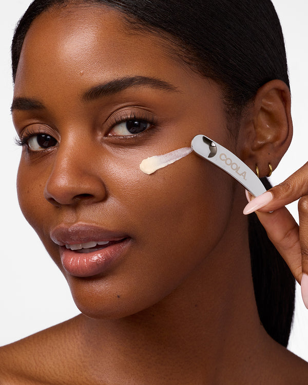 Close-up image of a woman applying eye cream with a metal applicator against a white background