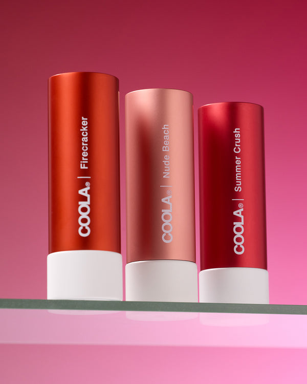 COOLA Mineral Tinted Liplux Firecracker, Nude Beach, and Summer Crush sitting on glass shelf against pink background.