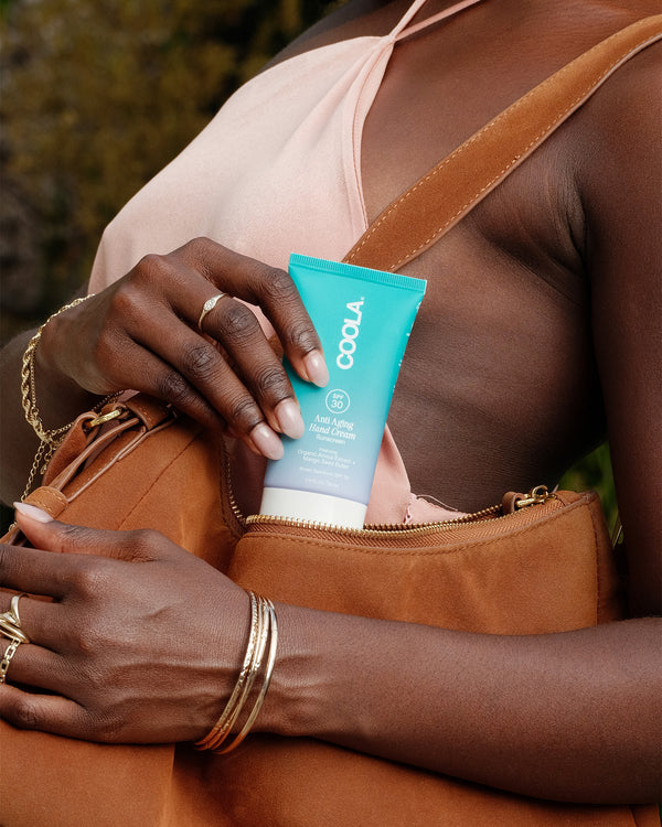 Person holding a turquoise tube of COOLA Anti-Aging Hand Cream with a tan leather bag, wearing gold bracelets and rings, featuring organic, premium skincare with SPF protection.
