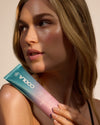 Dew Good Body Melt Lotion held against a womans shoulder in warm sunlight with glowing skin and a soft teal to pink package design