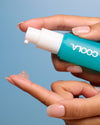 Hand holding a teal COOLA sunscreen bottle against a blue background