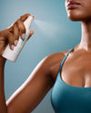 Person using a spray bottle labeled 'Coola' on their arm against a blue background