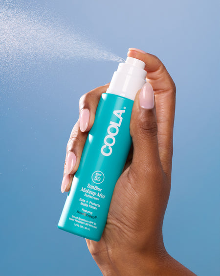 Coola sunscreen mist being sprayed by a hand against a blue background