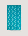 The COOLA x Business & Pleasure Beach Towel in a vibrant turquoise shade with a wavy pattern and fringe edges, perfect for relaxing days with organic, premium skincare essentials.