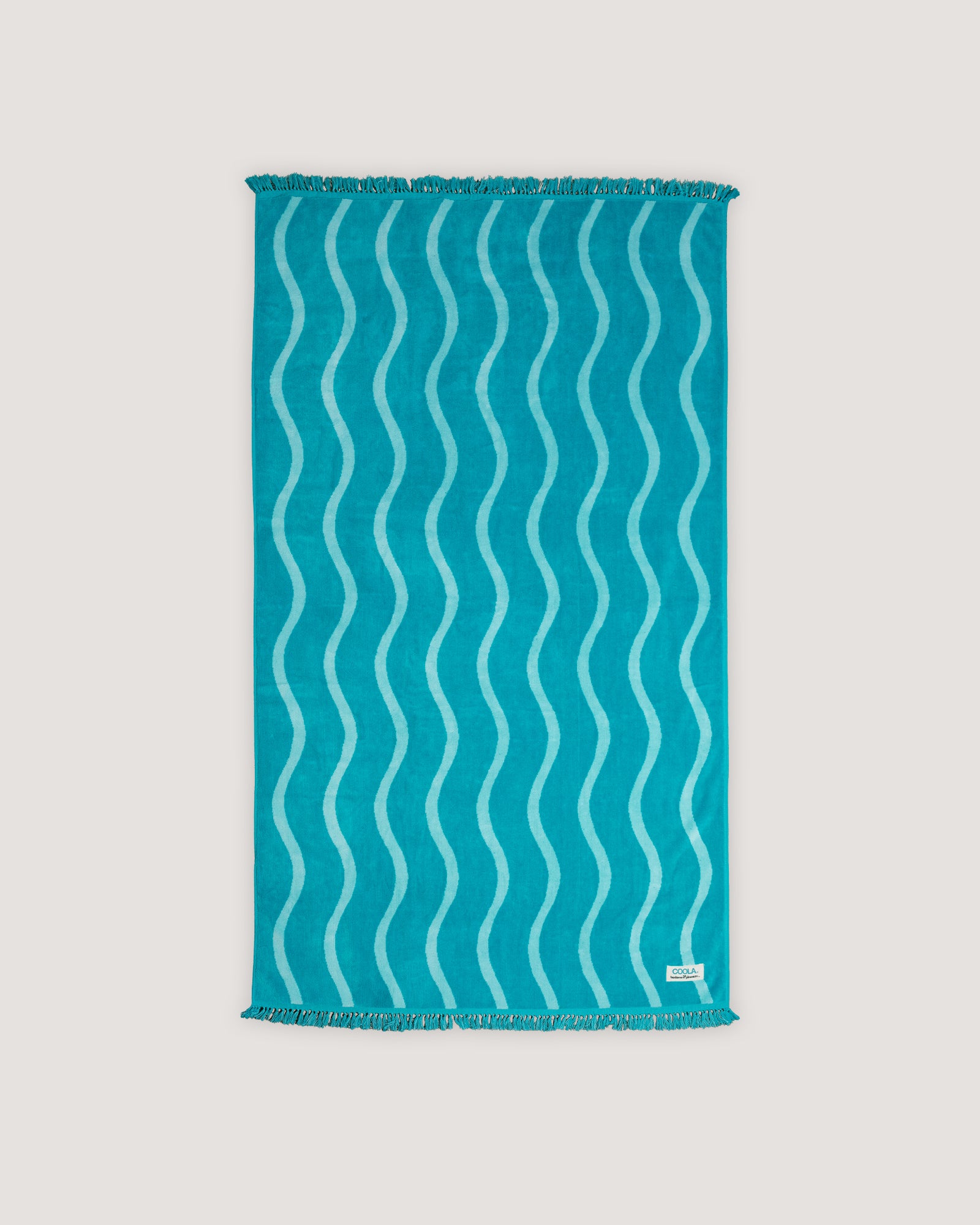 The COOLA x Business & Pleasure Beach Towel in a vibrant turquoise shade with a wavy pattern and fringe edges, perfect for relaxing days with organic, premium skincare essentials.