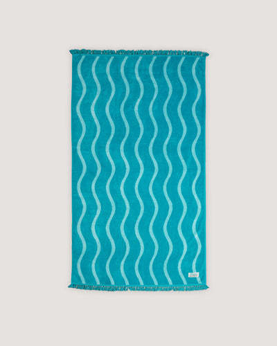 The COOLA x Business & Pleasure Beach Towel in a vibrant turquoise shade with a wavy pattern and fringe edges, perfect for relaxing days with organic, premium skincare essentials.