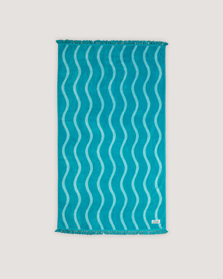 The COOLA x Business & Pleasure Beach Towel in a vibrant turquoise shade with a wavy pattern and fringe edges, perfect for relaxing days with organic, premium skincare essentials.