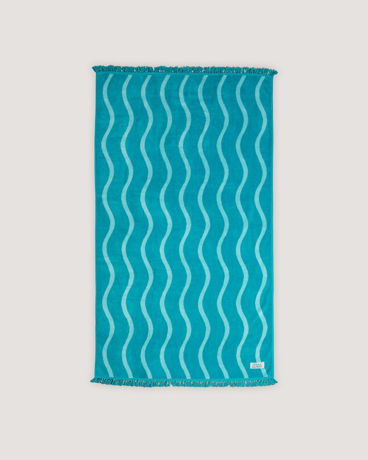 The COOLA x Business & Pleasure Beach Towel in a vibrant turquoise shade with a wavy pattern and fringe edges, perfect for relaxing days with organic, premium skincare essentials.