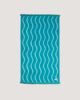 The COOLA x Business & Pleasure Beach Towel in a vibrant turquoise shade with a wavy pattern and fringe edges, perfect for relaxing days with organic, premium skincare essentials.