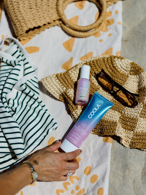 A hand reaches for COOLA sunscreen products lying on a patterned towel next to a woven hat and sunglasses, with a striped garment partially visible on the side.