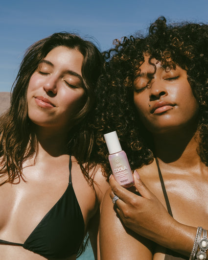 Two women with glowing skin wearing black swimsuits enjoy the sun, one holding a bottle of COOLA sunscreen, highlighting a natural and radiant sun care moment.