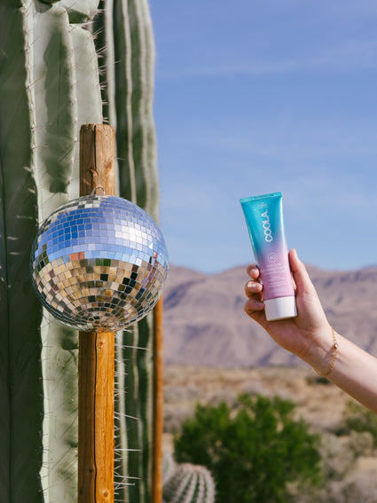 Hand holding a tube of COOLA sunscreen with a desert background featuring a tall cactus and a reflective disco ball hanging from a wooden post.