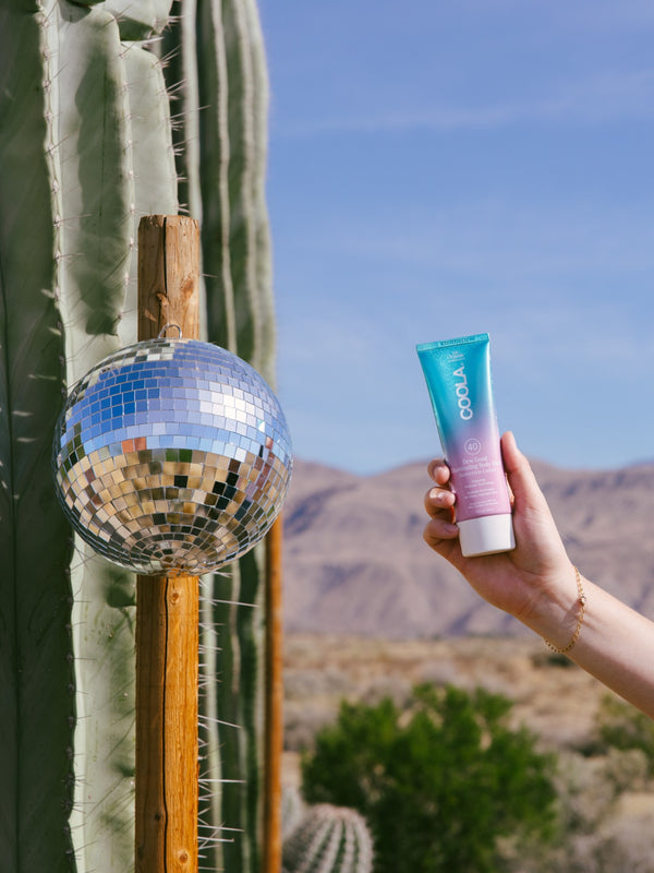 Hand holding a tube of COOLA sunscreen with a desert background featuring a tall cactus and a reflective disco ball hanging from a wooden post.