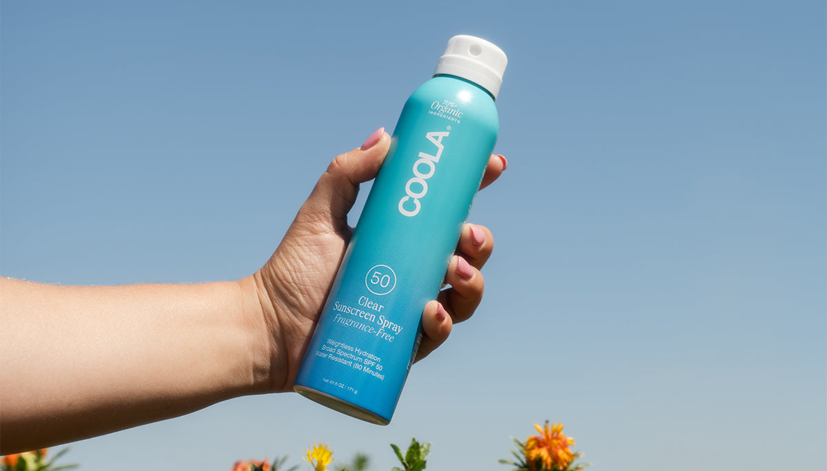 Hand holding COOLA Clear Sunscreen Spray Fragrance-Free SPF 50 with blue sky background.