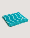 The COOLA x Business & Pleasure Beach Towel folded with a turquoise and light blue wavy pattern and fringed edges, showcasing casual, beach-ready style.