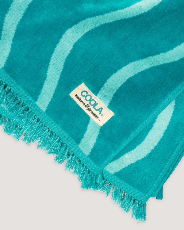 Close-up of a turquoise and light blue striped towel with fringed edges featuring a sewn label that reads The COOLA x Business & Pleasure Beach Towel, perfect for organic, premium skincare lovers enjoying feel-good sunscreen days.