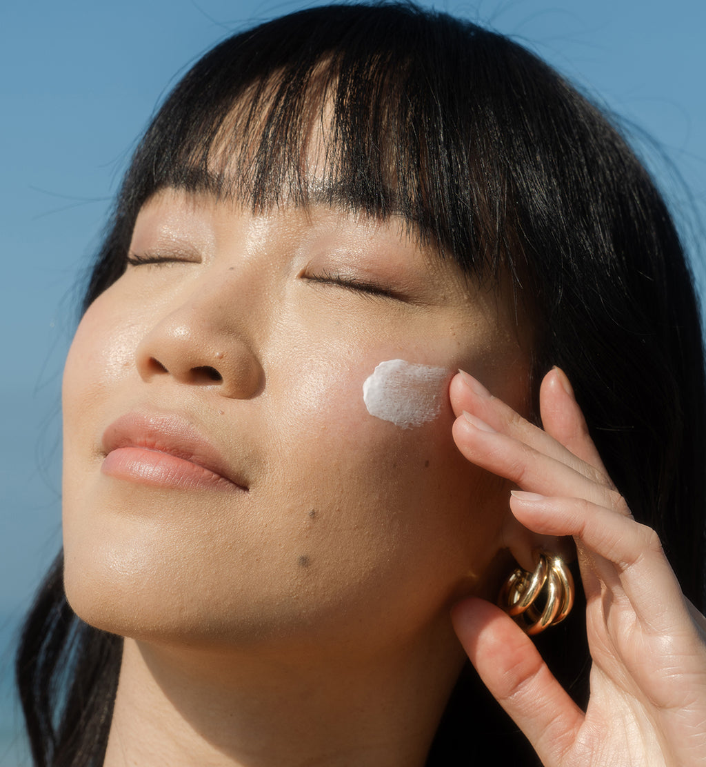 Close-up of woman applying sunscreen lotion to cheek with eyes closed and blue sky in the background