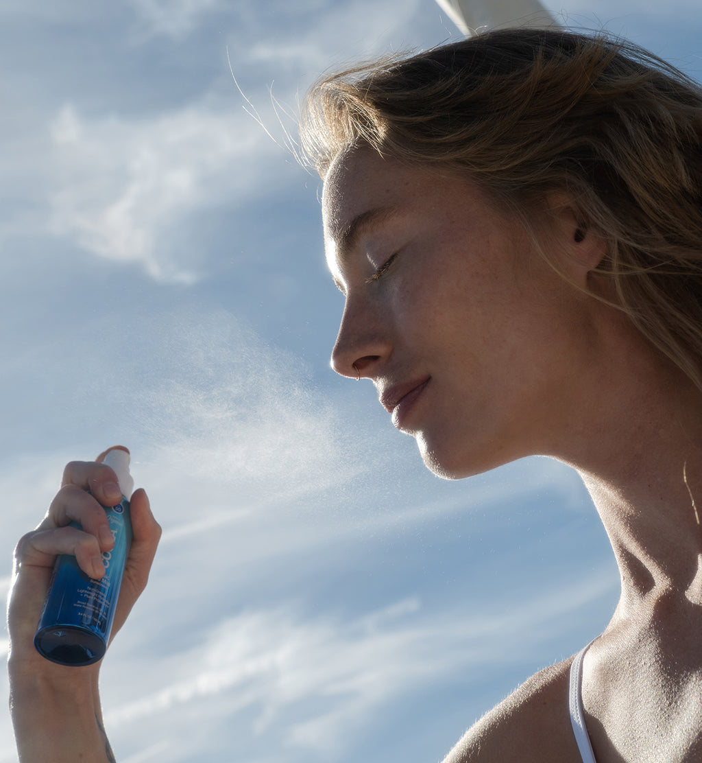 Woman applying COOLA Classic Face Mist Sunscreen to face with eyes closed and blue sky in background
